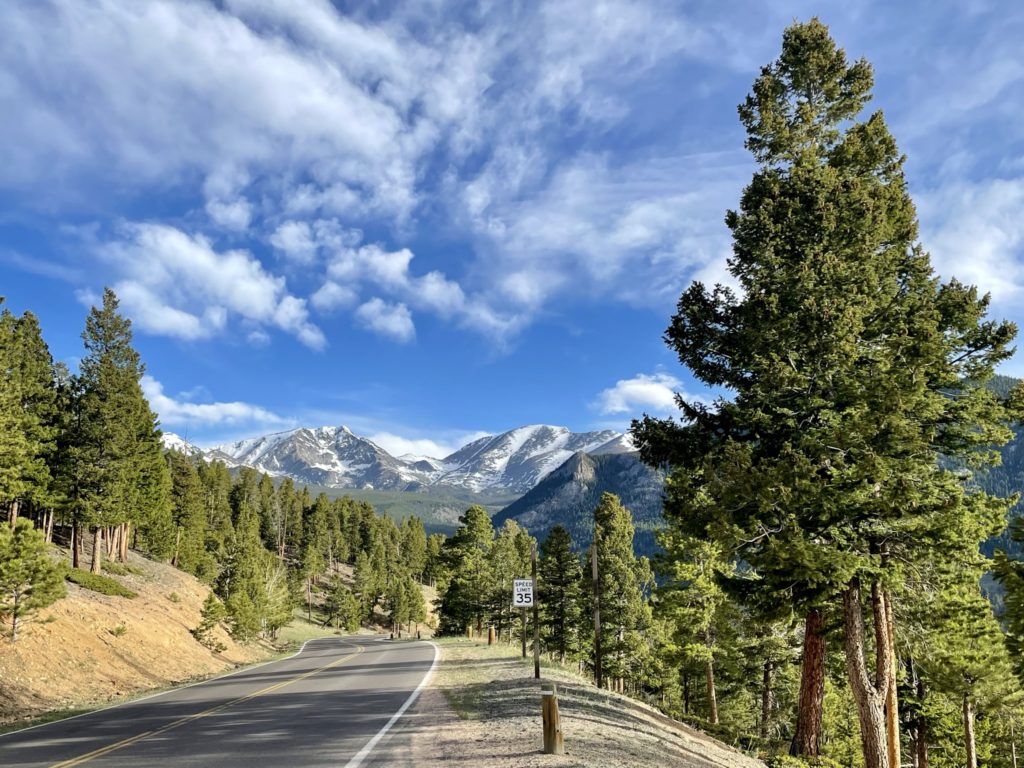 Rocky Mountain National Park road twists and turns into snow capped mountains with green trees and light clouds in blue sky