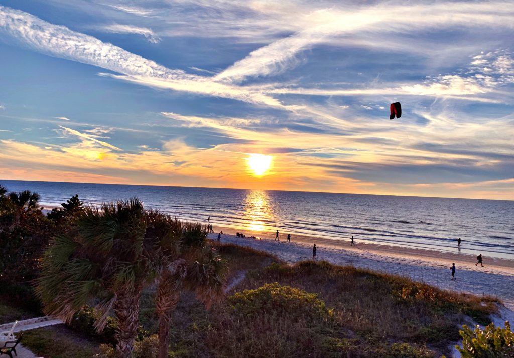 View of sun setting over the blue ocean, with view of beach, a kite, and clouds in a blue sky