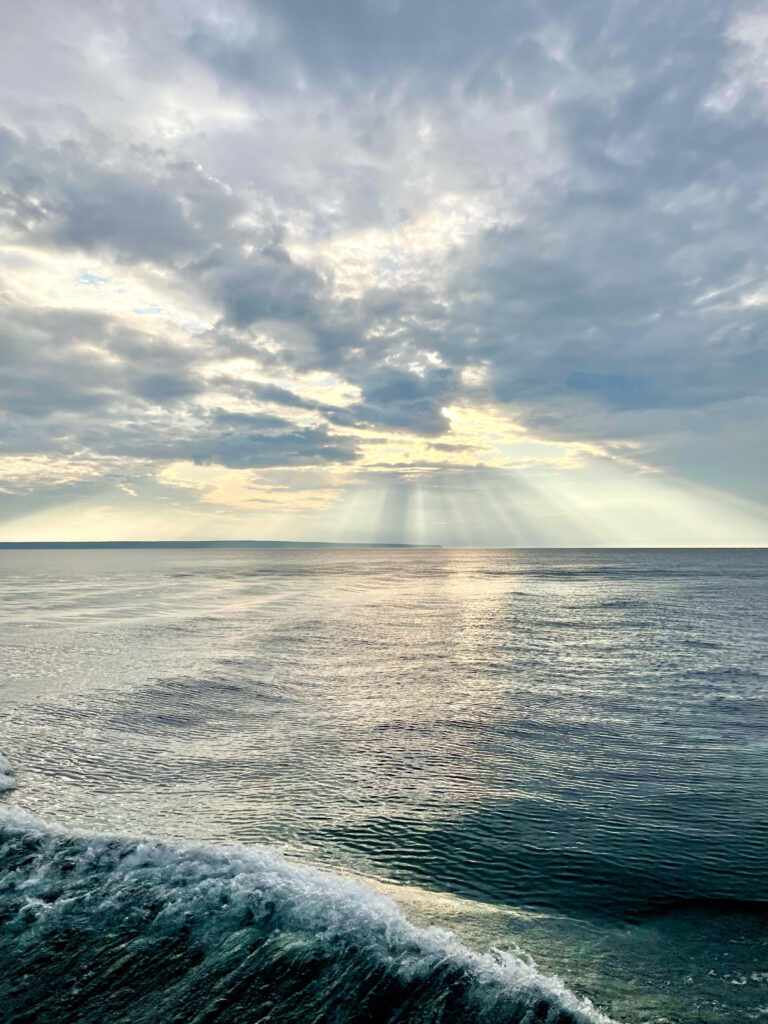 View of blue green lake with waves in the corner, looking out to the blue gray clouds as rays of sun shine through the clouds
