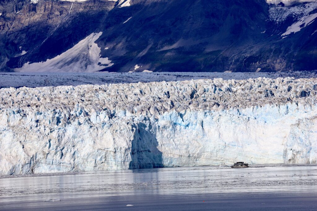 Close up of Hubbard Glacier with blue, white, and black striations and mountains in background and a tiny catamaran in front of the glacier on the water