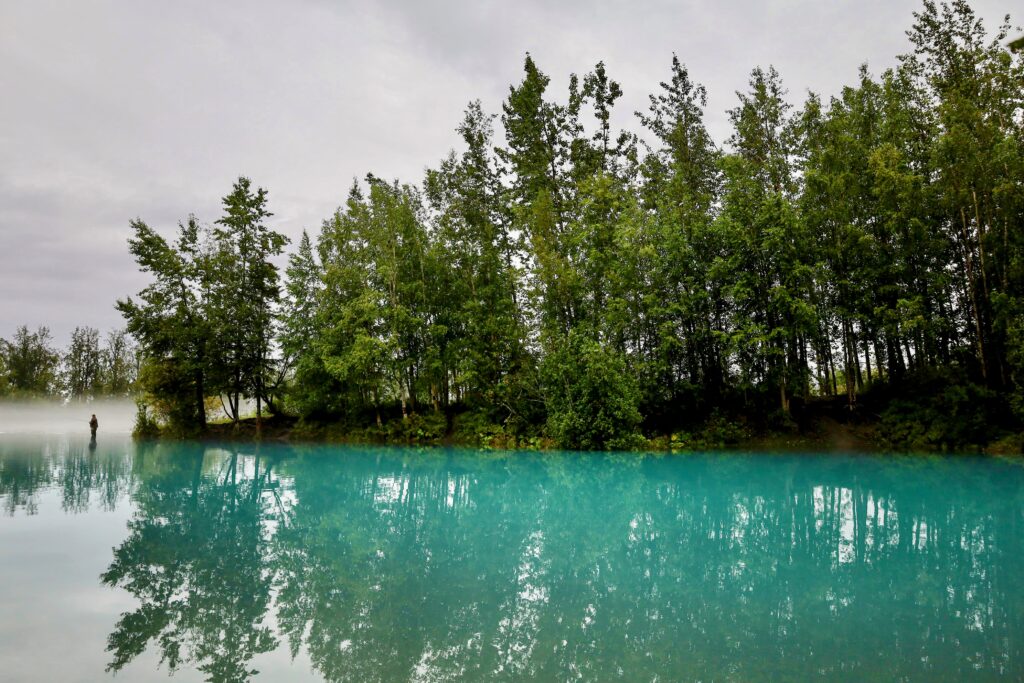 Tiny fisherman fishing in turquoise water along shore with tall green trees