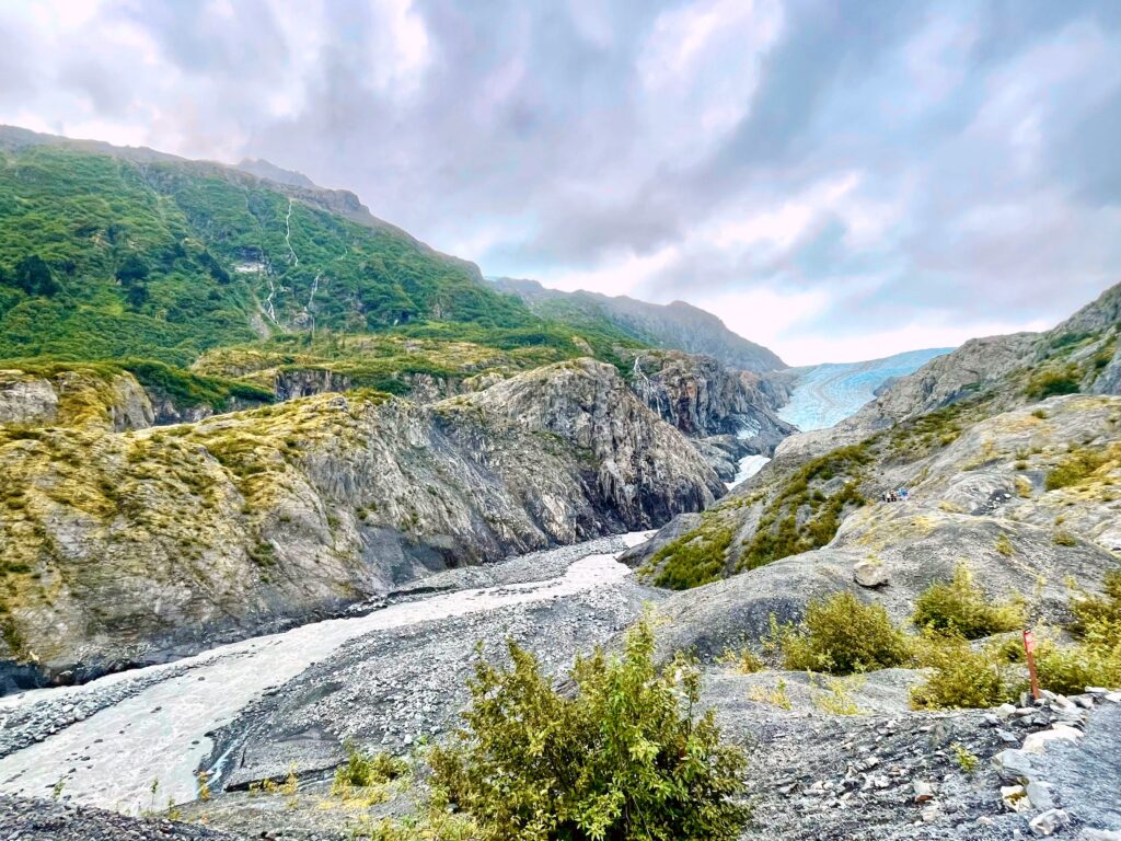 Exit Glacier with lush green mountains, a stream below the glacier, and a rocky mountain with grey clouds