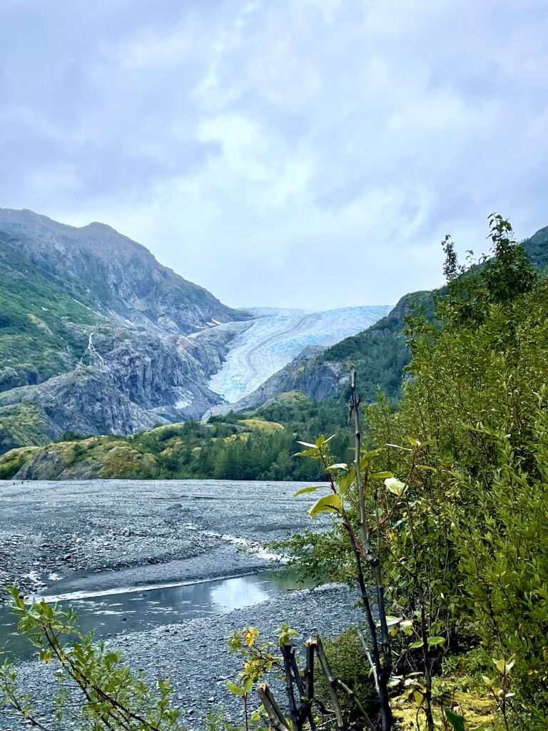 Exit glacier between two mountains with stream, rocky shore, gray clouds, and greenery