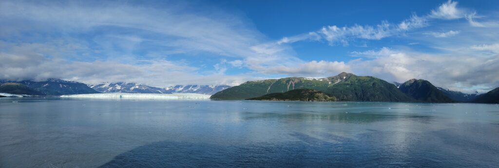 Pano of Hubbard Glacier with water, glacier, green mountains, and blue skies with puffy clouds