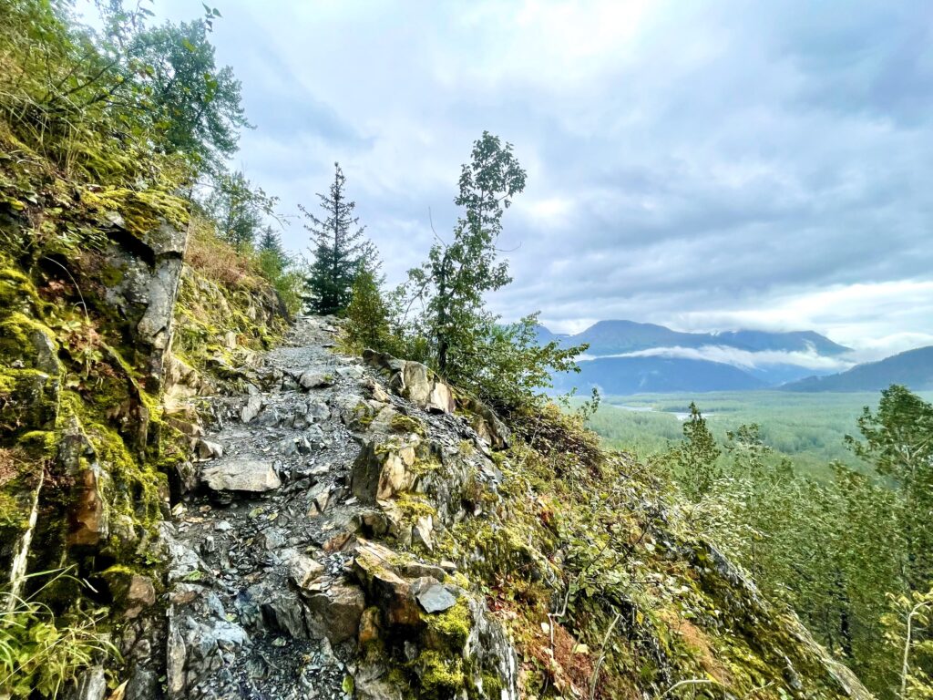 Steep rocky trail overlooks green valley with a river and mountains with clouds in the background