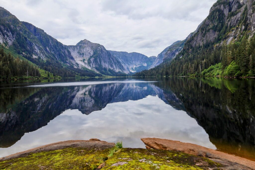 View of lake with dramatic blue/grey mountains on either side and reflecting on water with green trees and shrubs