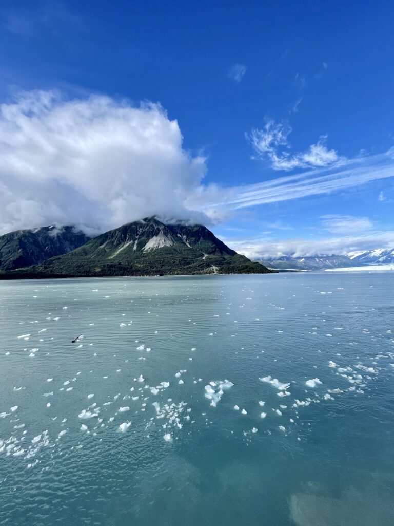 Ice chunks in blue water with dramatic green mountains, a white glacier, and puffy clouds in blue sky