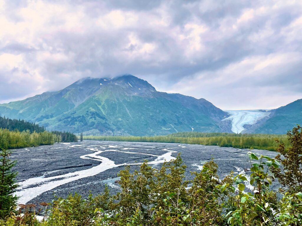 Cloudy gray sky with green mountain and water running in different areas through a rocky riverbed with a glacier in the background