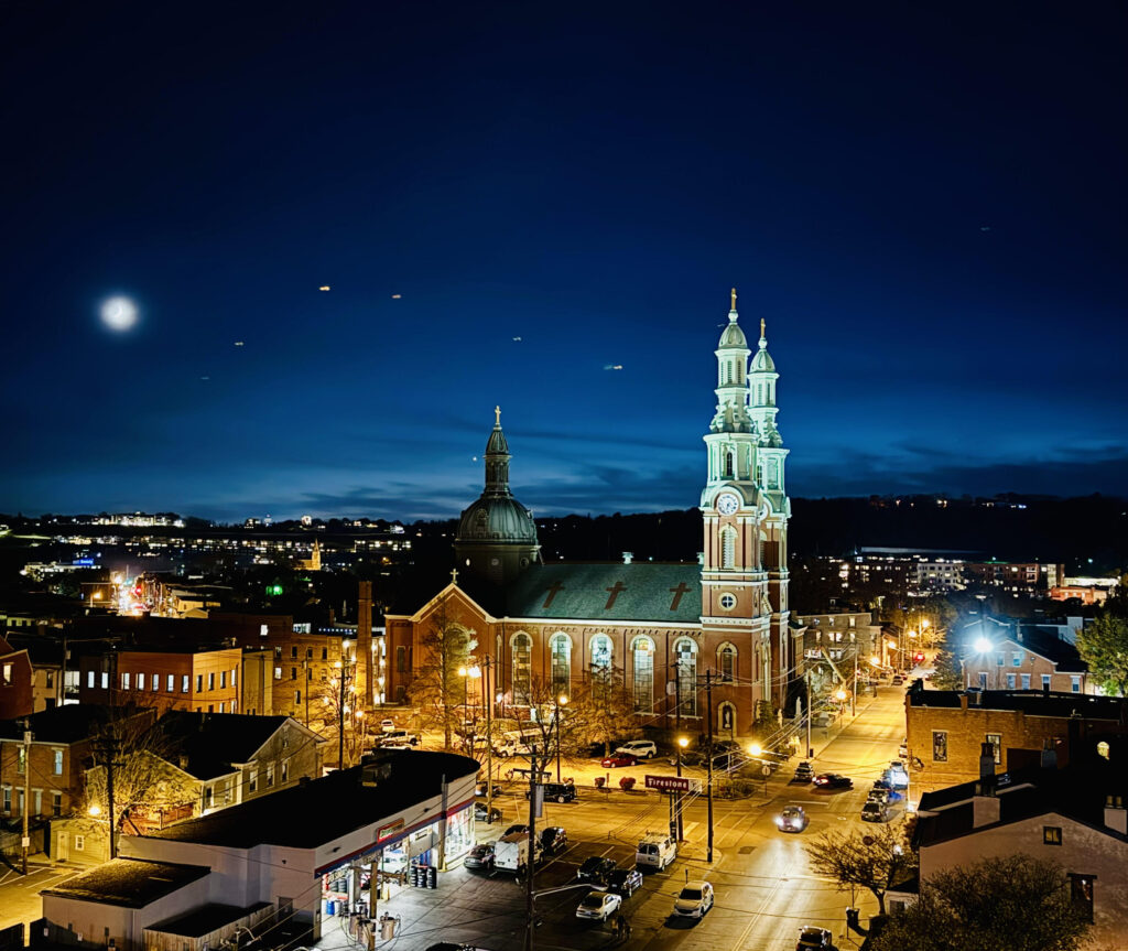 Photo of dark blue night sky with fuzzy moon and lights on a large church with cars on the street