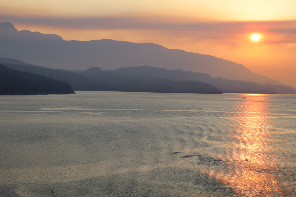 Sun setting over land with hazy orange and purple sky and bluish reflection in the water, land is varying shades of purple.