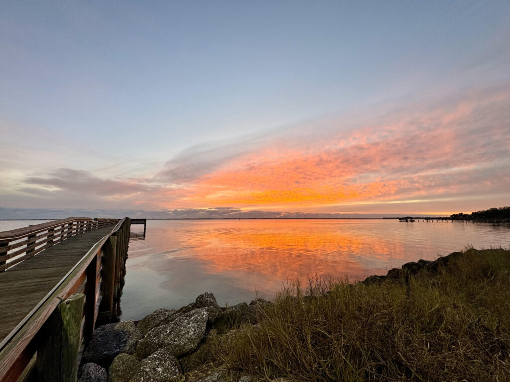 view of orange sunrise over water and a long pier into water with rocks along the shore