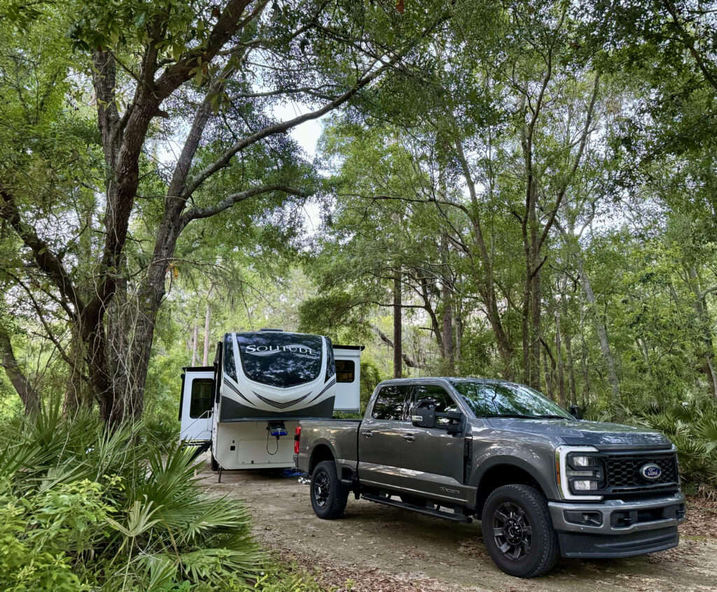 Gray truck and black and white fifth wheel camper tucked into green trees