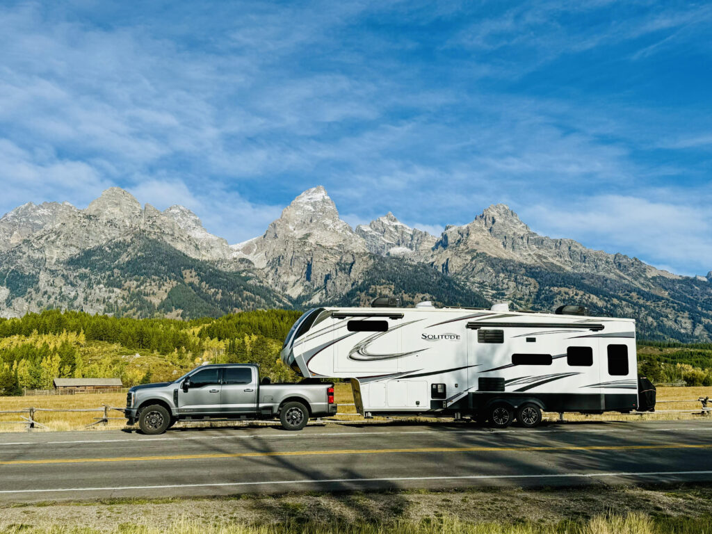 View of grey truck pulling white trailer on road in front of the Grand Teton mountains with blue sky