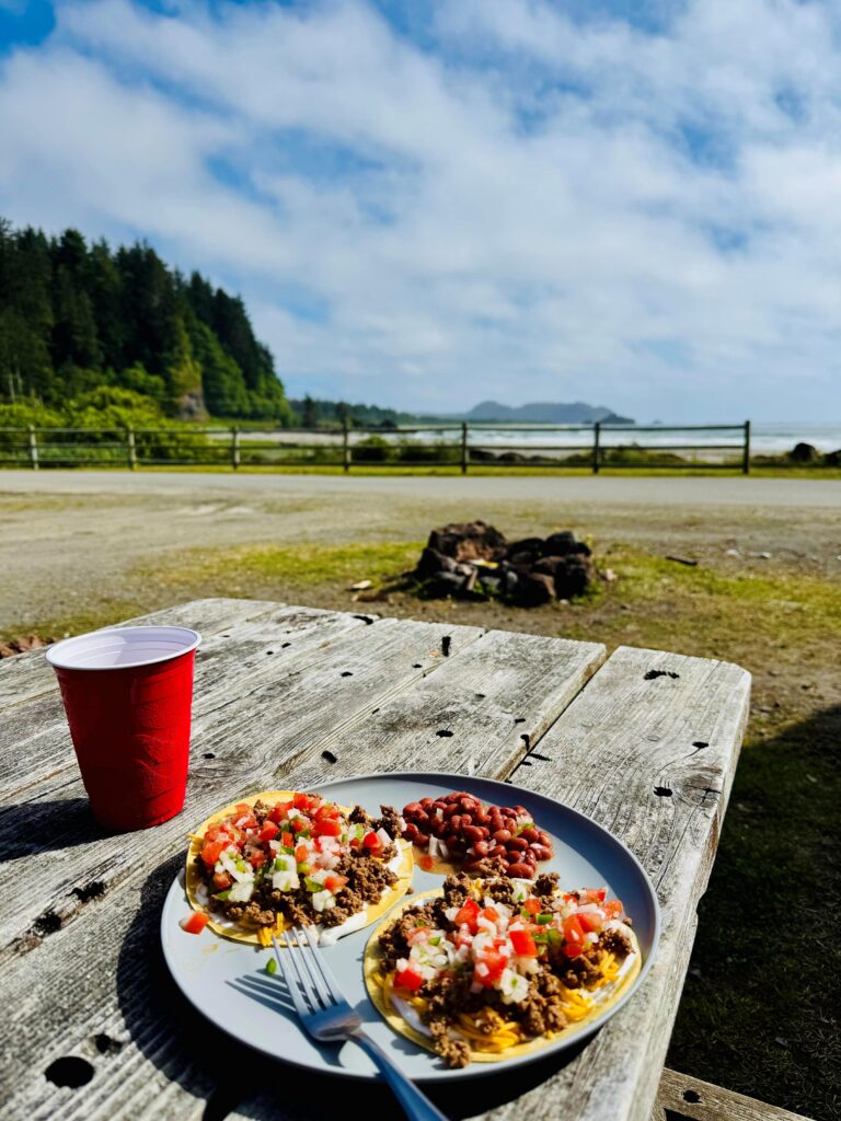 Picture of a plate of tacos and beans on a picnic table with a red cup and the ocean and trees in the background