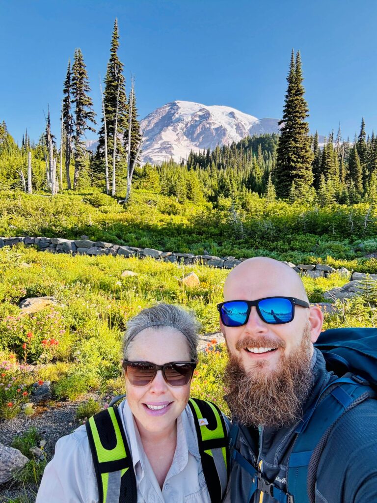 Picture of a man and woman in hiking gear with Mt Rainier in the background and green fields and trees
