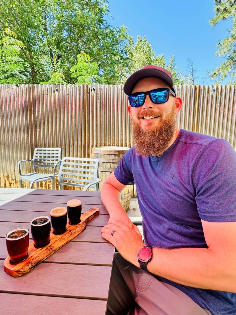 Man with reddish beard, hat, and sunglasses in front of a flight of beer on an outside table.