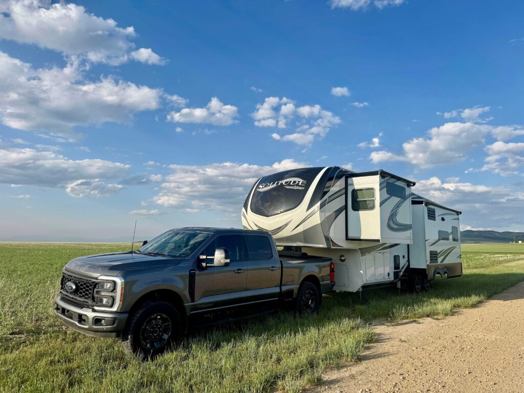 Large gray truck with large white and black RV pulled over along a dirt road in a green field with blue skies and puffy clouds